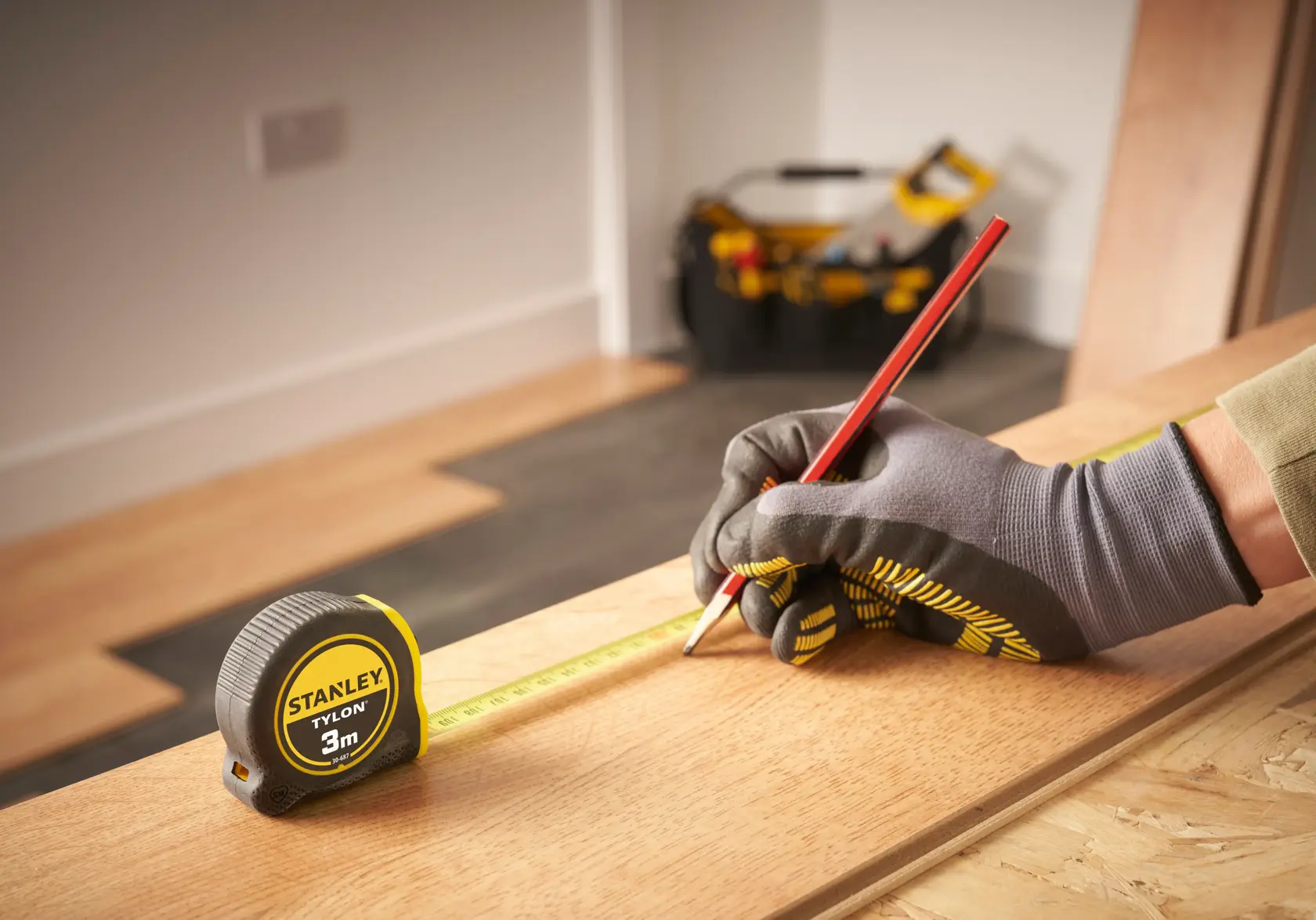 Foto des STANLEY 0-30-687 Maßbands auf Holz, Arbeiter mit Handschuhen markiert mit rotem Stift, Werkzeugtasche im Hintergrund.