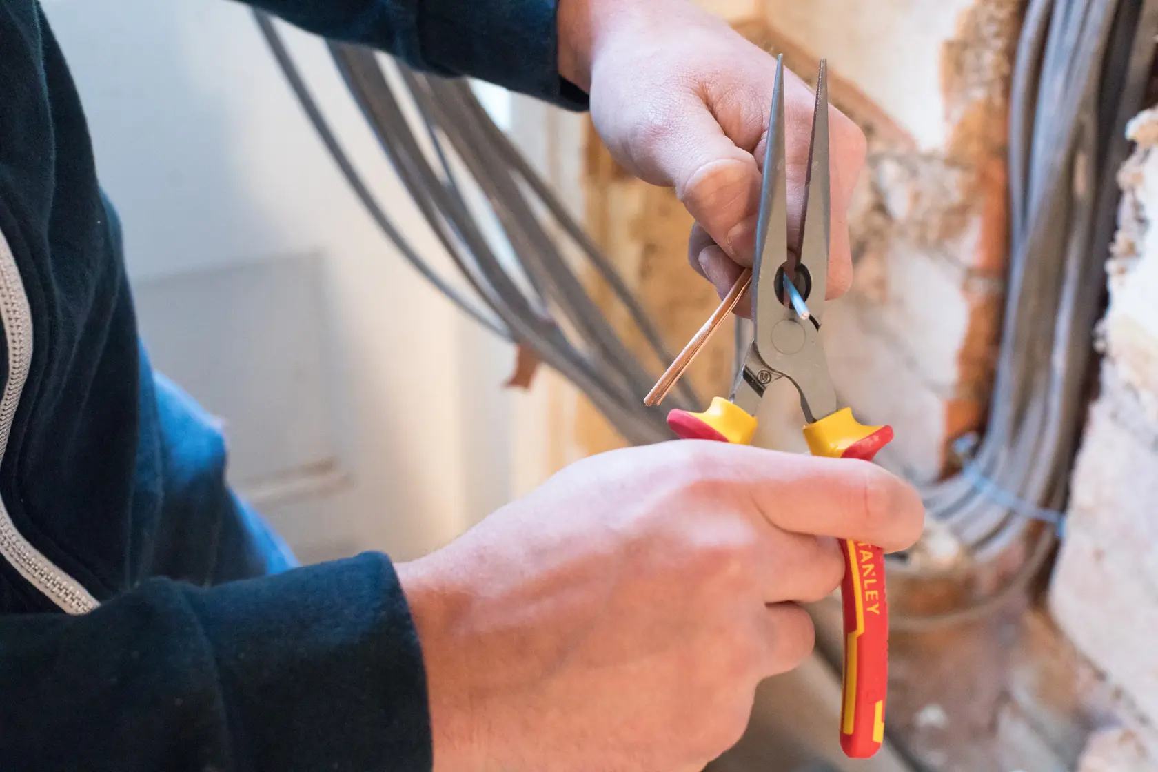 STANLEY 0-84-007 Langnasenzange in der Hand, greift Draht nahe Wand, Werkzeug mit gelben und roten Griffen.