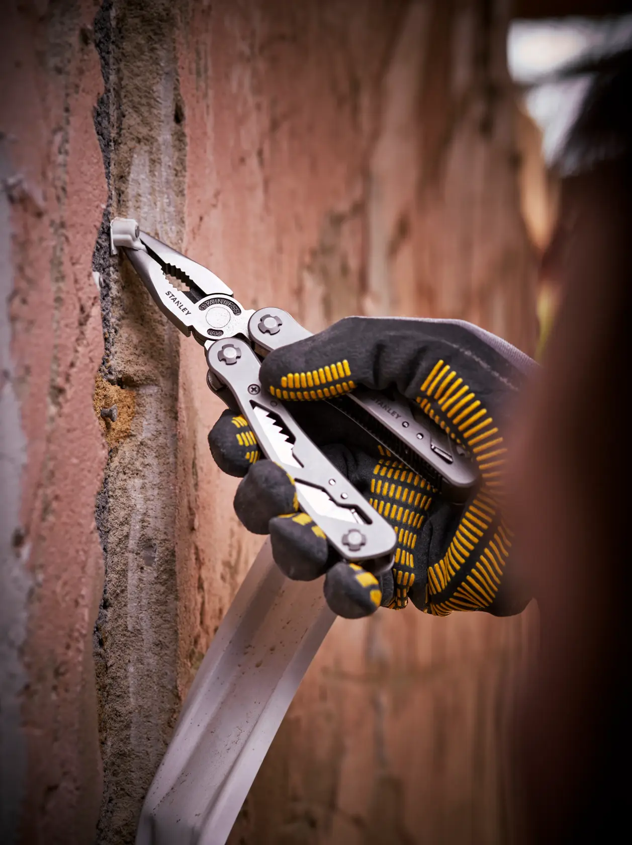 STANLEY 0-84-519 Multitool-Zange an einer Ziegelwand, Nahaufnahme-Fotografie einer behandschuhten Hand.