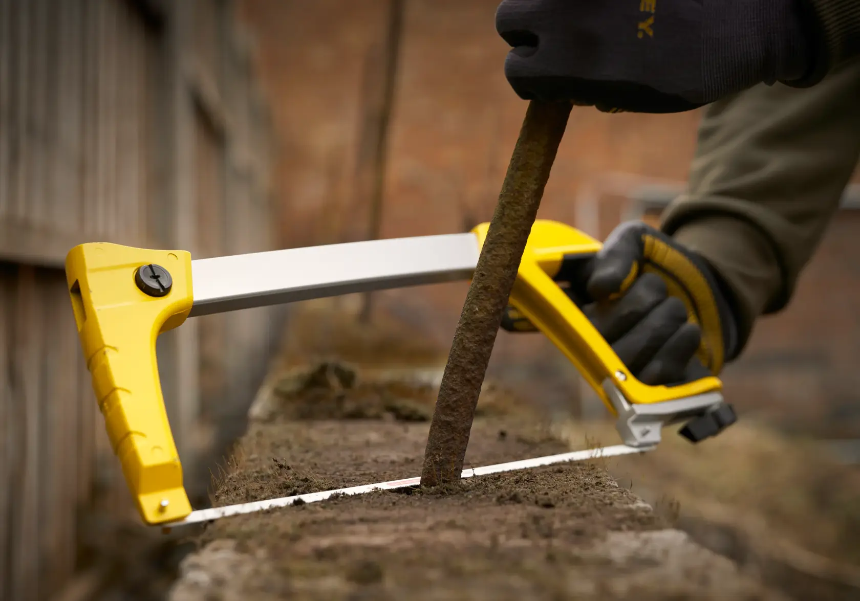 STANLEY 1-20-110 Säge im Einsatz, schneidet eine rostige Metallstange im Freien, Nahaufnahme des gelben Rahmens und der behandschuhten Hand.