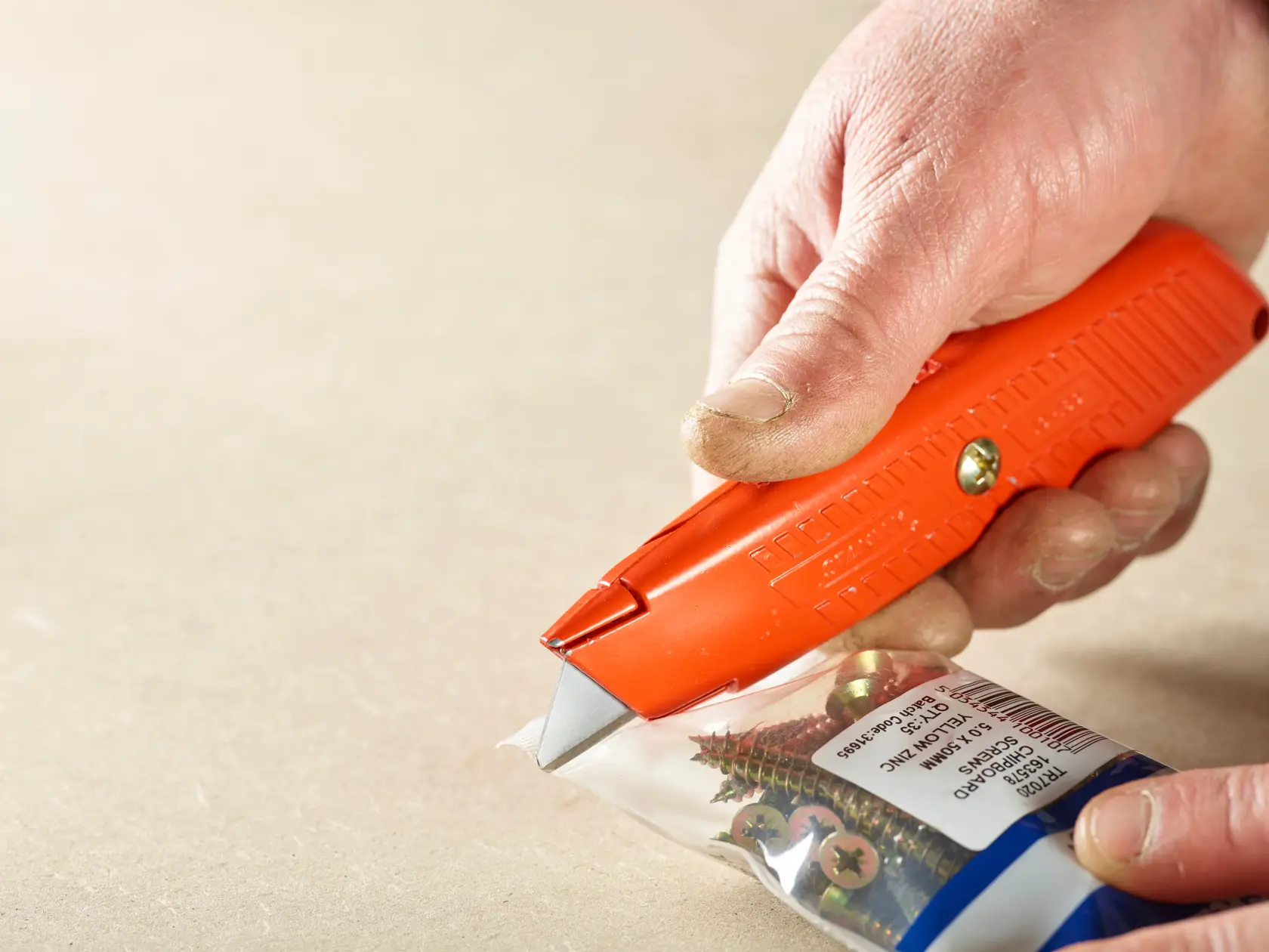STANLEY 2-11-987 Universalmesser schneidet Kunststoffverpackung mit Schrauben, Nahaufnahme von orangem Griff und Klinge.