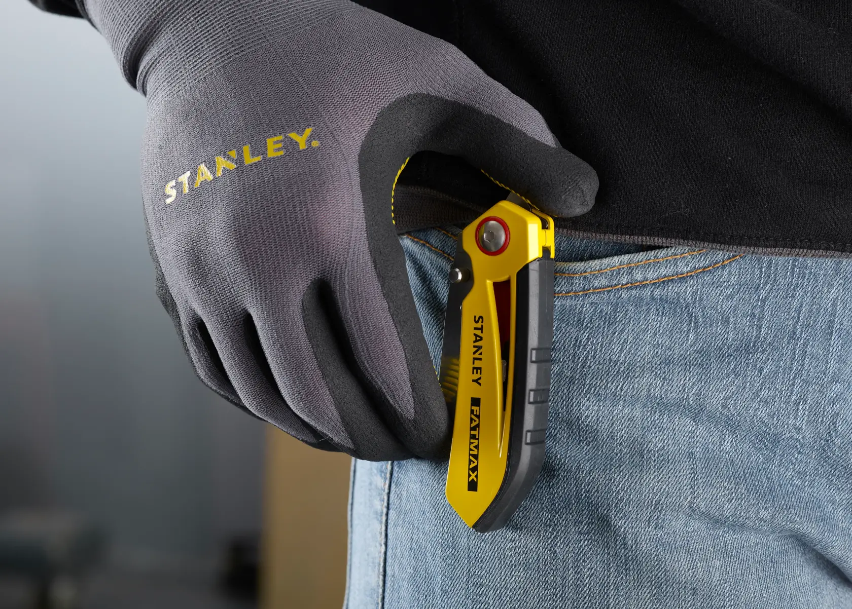 STANLEY FMHT0-10827 klappbares Universalmesser in Gelb und Schwarz am Jeans befestigt, behandschuhte Hand hält es.