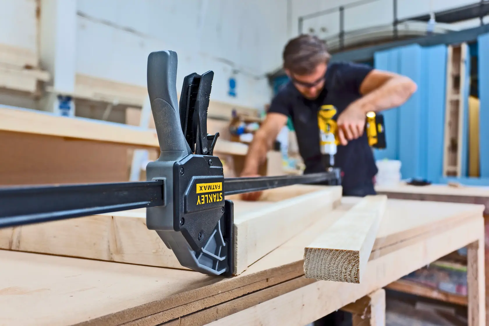 STANLEY FMHT0-83240 Zwinge hält Holz auf Werkbank, Werkstatt im Hintergrund, unscharfer Handwerker.