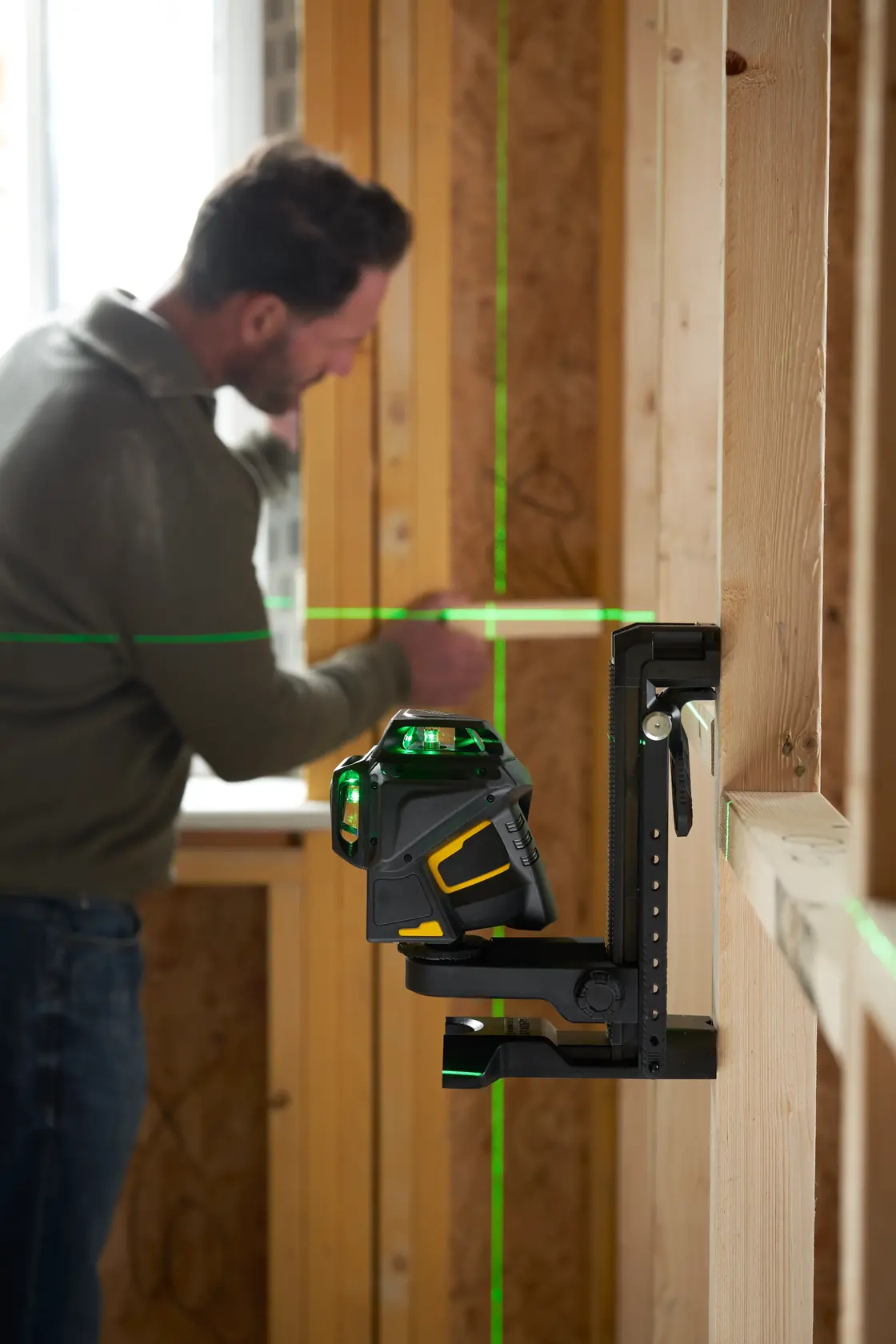 STANLEY FMHT1-77356 Laser-Nivelliergerät projiziert grüne Linien in Holzrahmenraum, Mann arbeitet hinten.