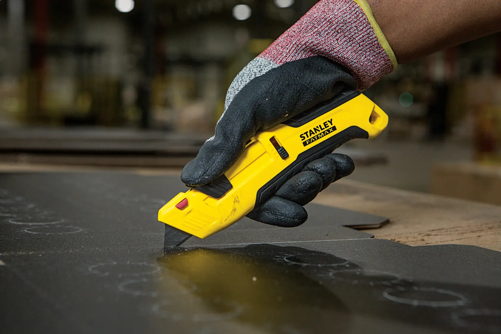 Foto des STANLEY FMHT10365-0 Cuttermessers in behandschuhter Hand, schneidet dunkles Material in Werkstatt.