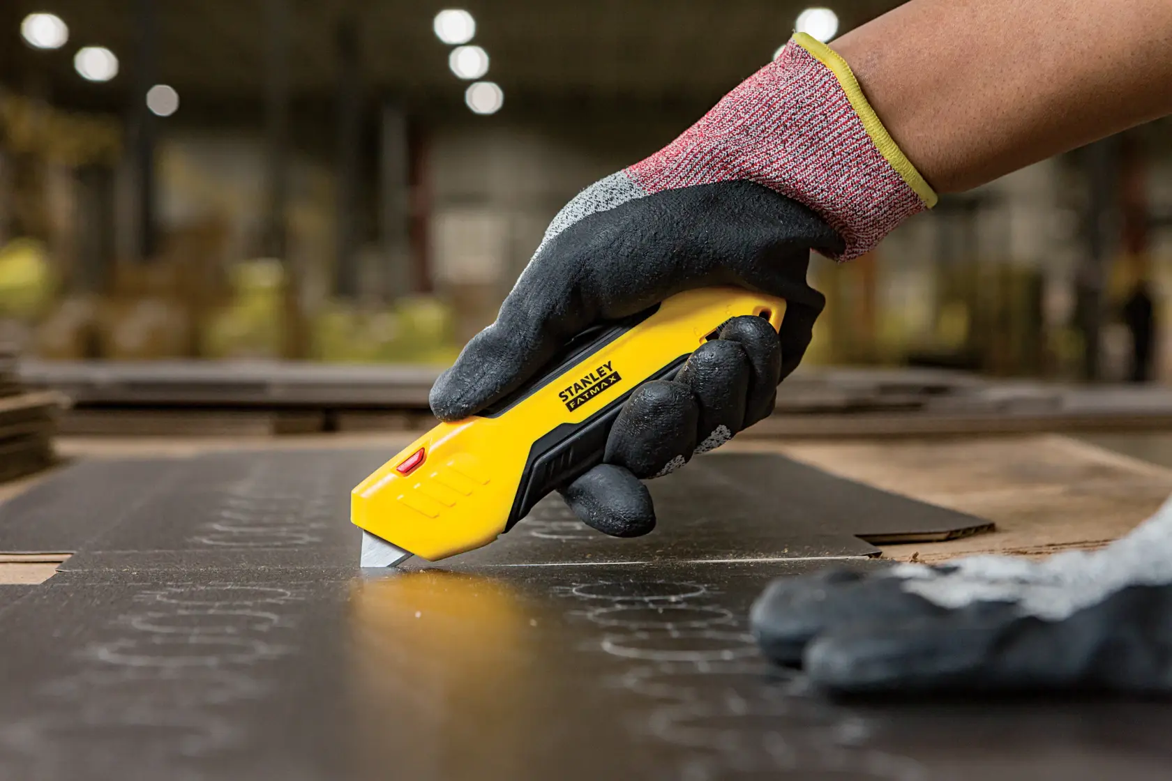Stanley FMHT10369-0 Messer schneidet Material, gehalten von einer behandschuhten Hand im Industrieumfeld.