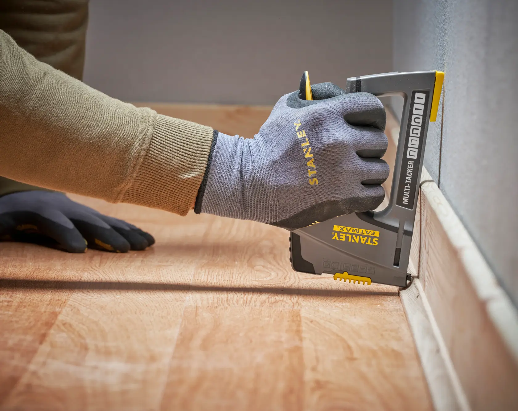 Foto einer Hand mit Stanley-Handschuhen, die einen STANLEY FMHT6-70868 Multitacker an einer Fußleiste verwendet, Seitenansicht.