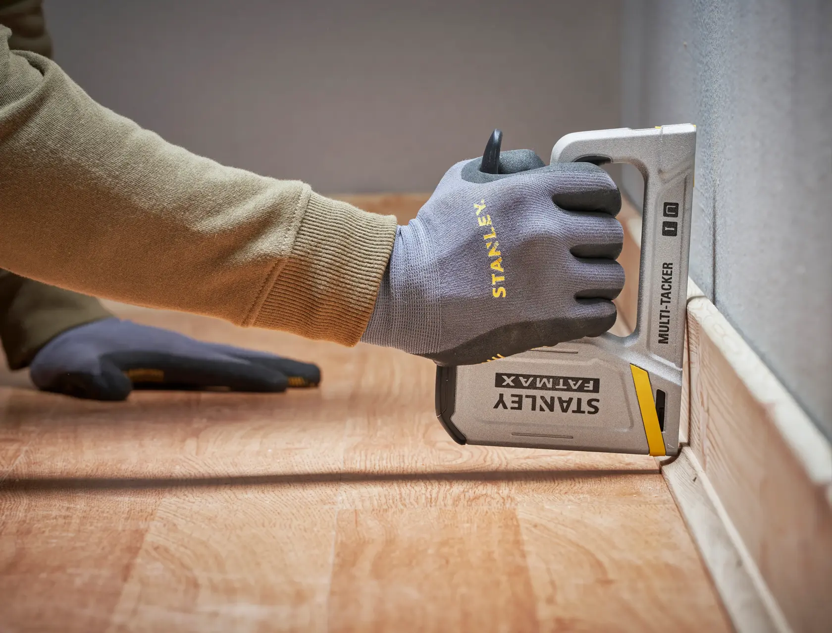 Foto einer behandschuhten Hand mit einem STANLEY FATMAX FMHT70443-0 Multi-Tacker auf dem Boden an einer Wand.