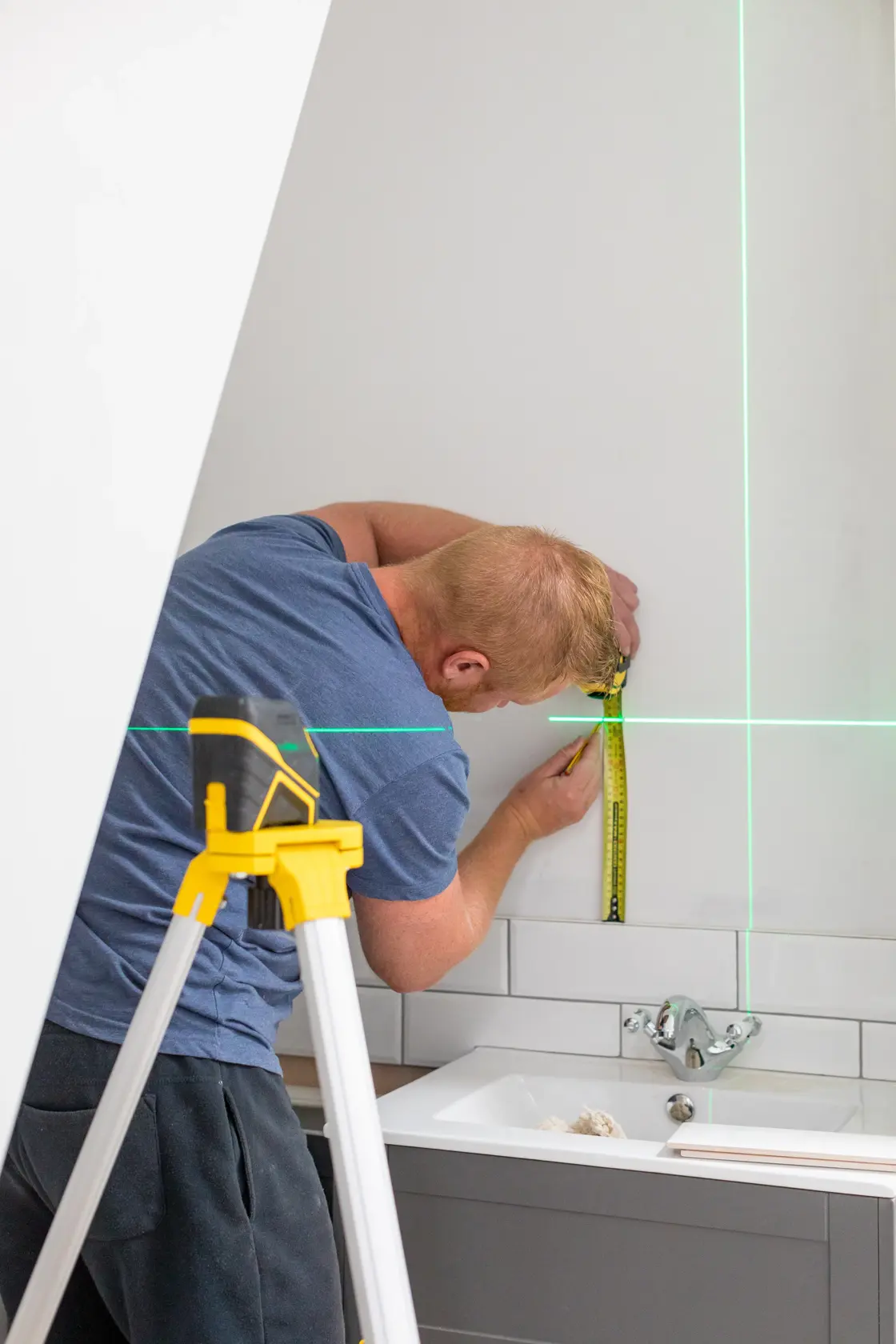STANLEY FMHT77586-1 Laser-Nivelliergerät auf Stativ projiziert grüne Linien, während ein Mann die Wand im Bad misst.