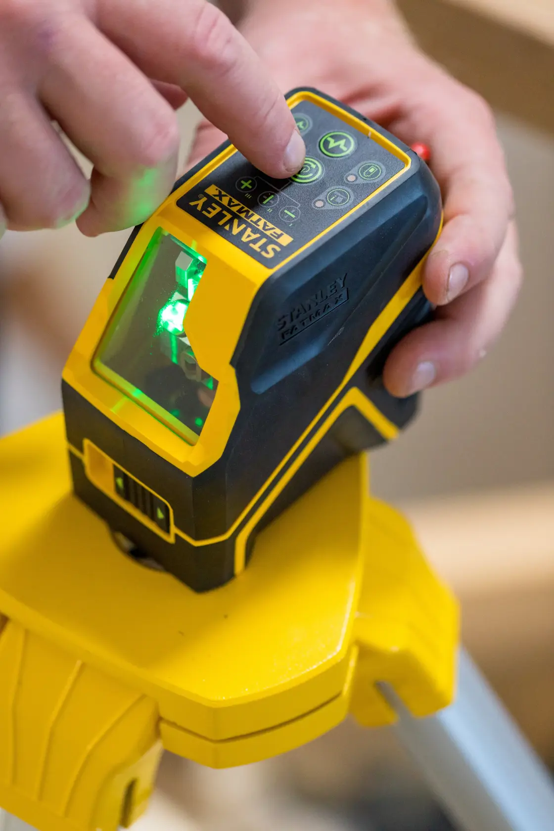 STANLEY FMHT77595-1 Laser-Nivelliergerät auf Stativ, Nahaufnahme einer Hand auf Bedientasten mit grünem Laser an.