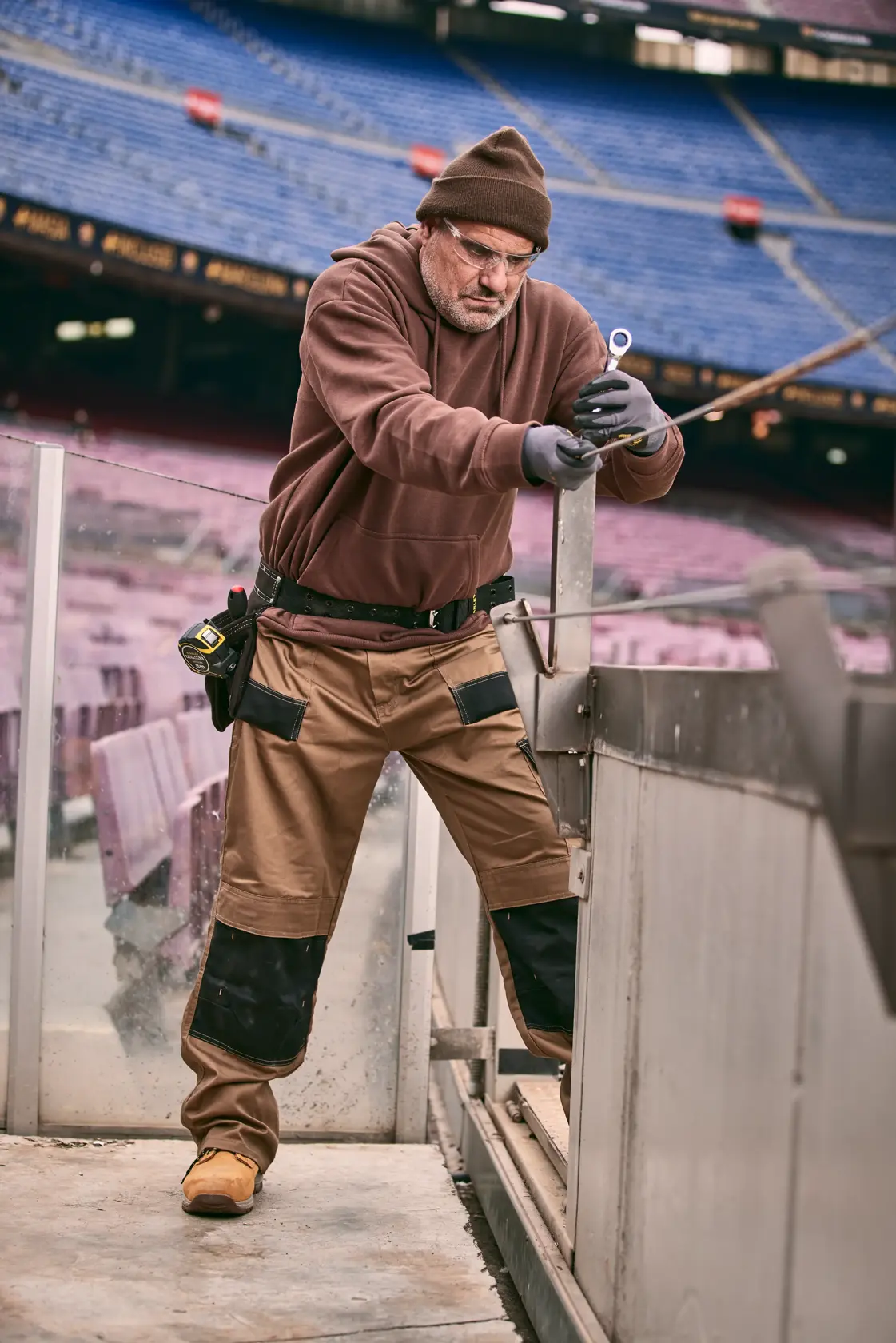 STANLEY FMMT82848-0 Fotografie eines Arbeiters in brauner Arbeitskleidung mit Handwerkzeug in einem leeren Stadion.