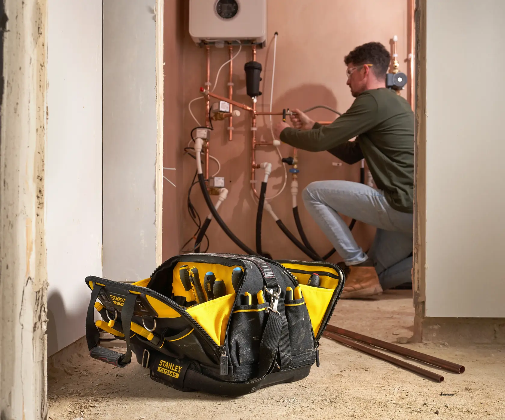 STANLEY FMST1-73607 Werkzeugtasche mit Werkzeugen am Boden, Arbeiter repariert Rohre im Hintergrund.