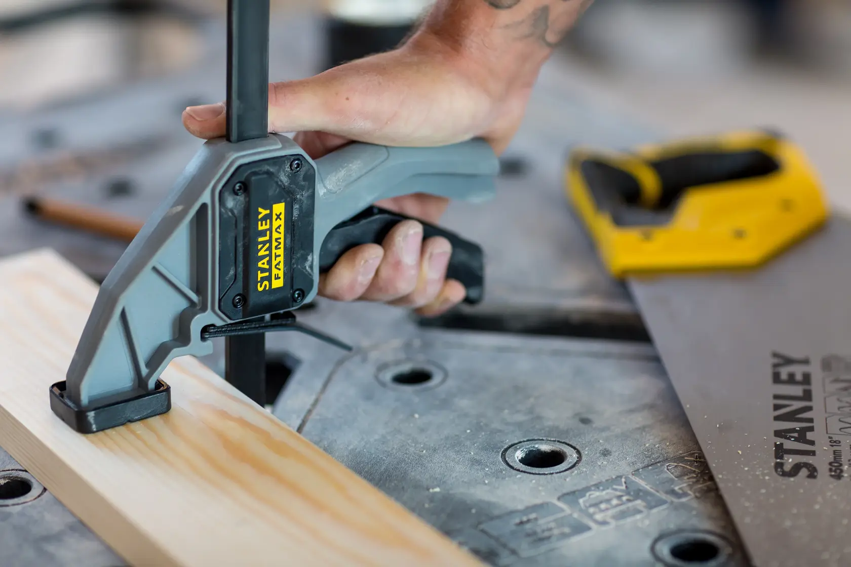 STANLEY FMST1-75672 Schraubzwinge fixiert Holz auf Werkbank, Nahaufnahme mit Hand am Griff, Säge im Hintergrund.