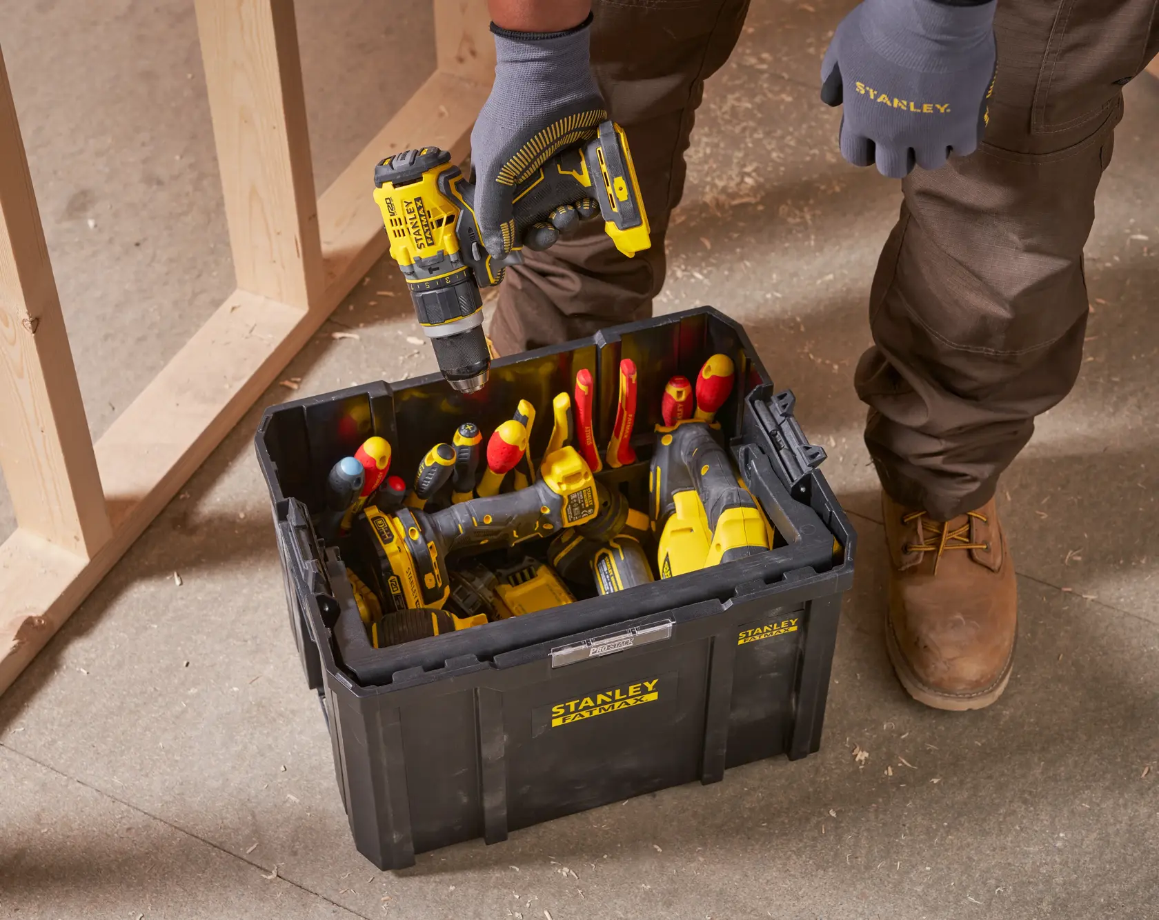 STANLEY FMST1-75794 schwarze Werkzeugbox auf Baustellenboden, gefüllt mit Hand- und Elektrowerkzeugen, Person hält Bohrmaschine.