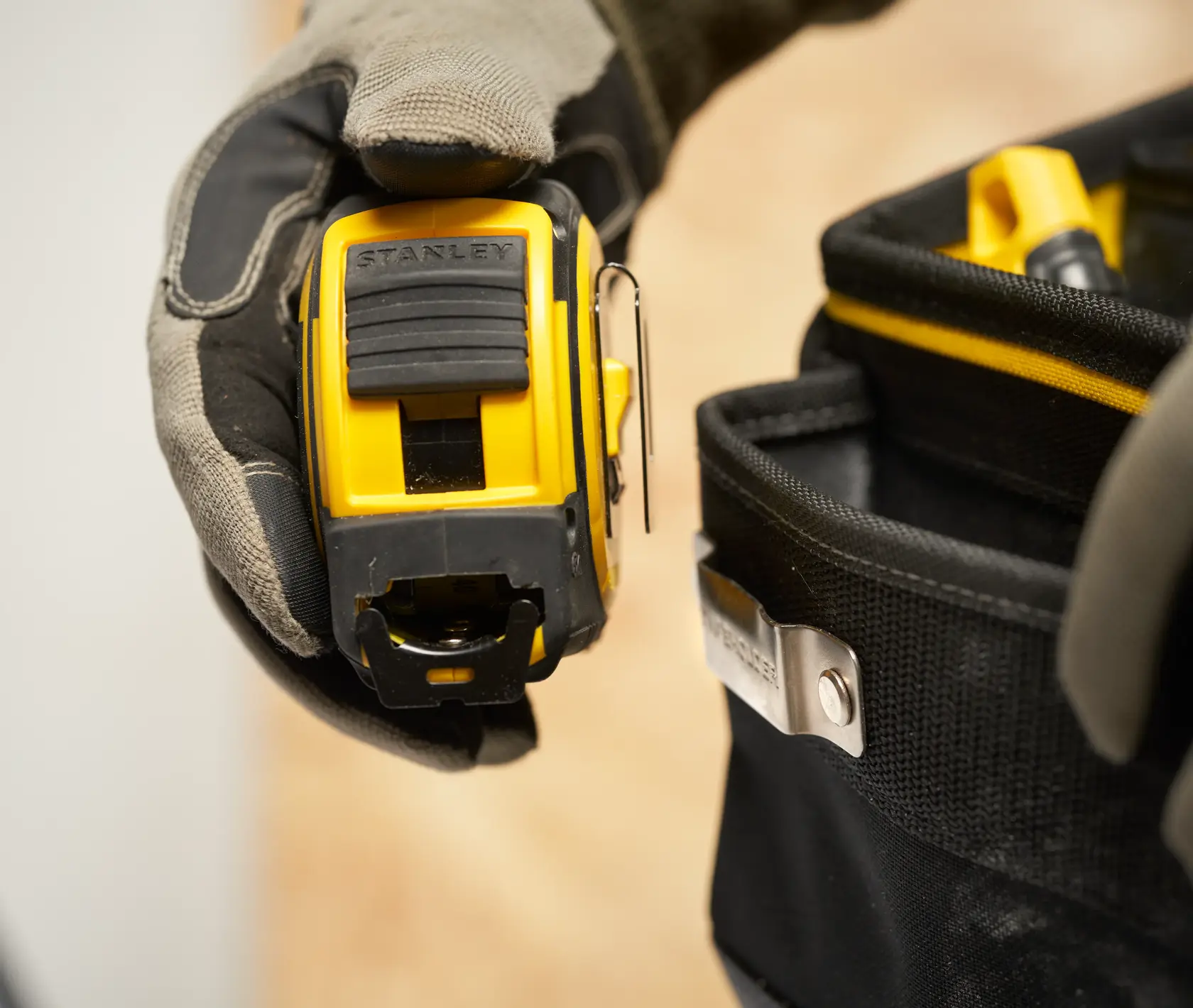 Foto vom STANLEY FMST17624-1 Maßband in behandschuhter Hand neben schwarzer Werkzeugtasche.