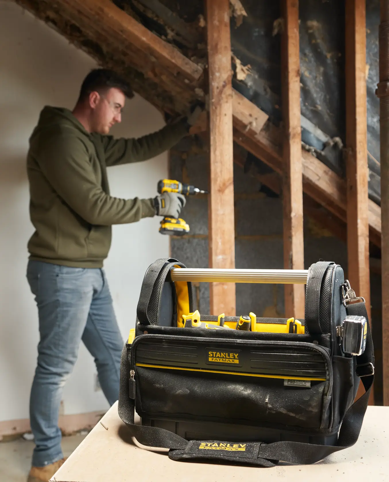 STANLEY FMST17626-1 Werkzeugtasche mit Griff und Schulterriemen auf Tisch, Mann arbeitet im Hintergrund.