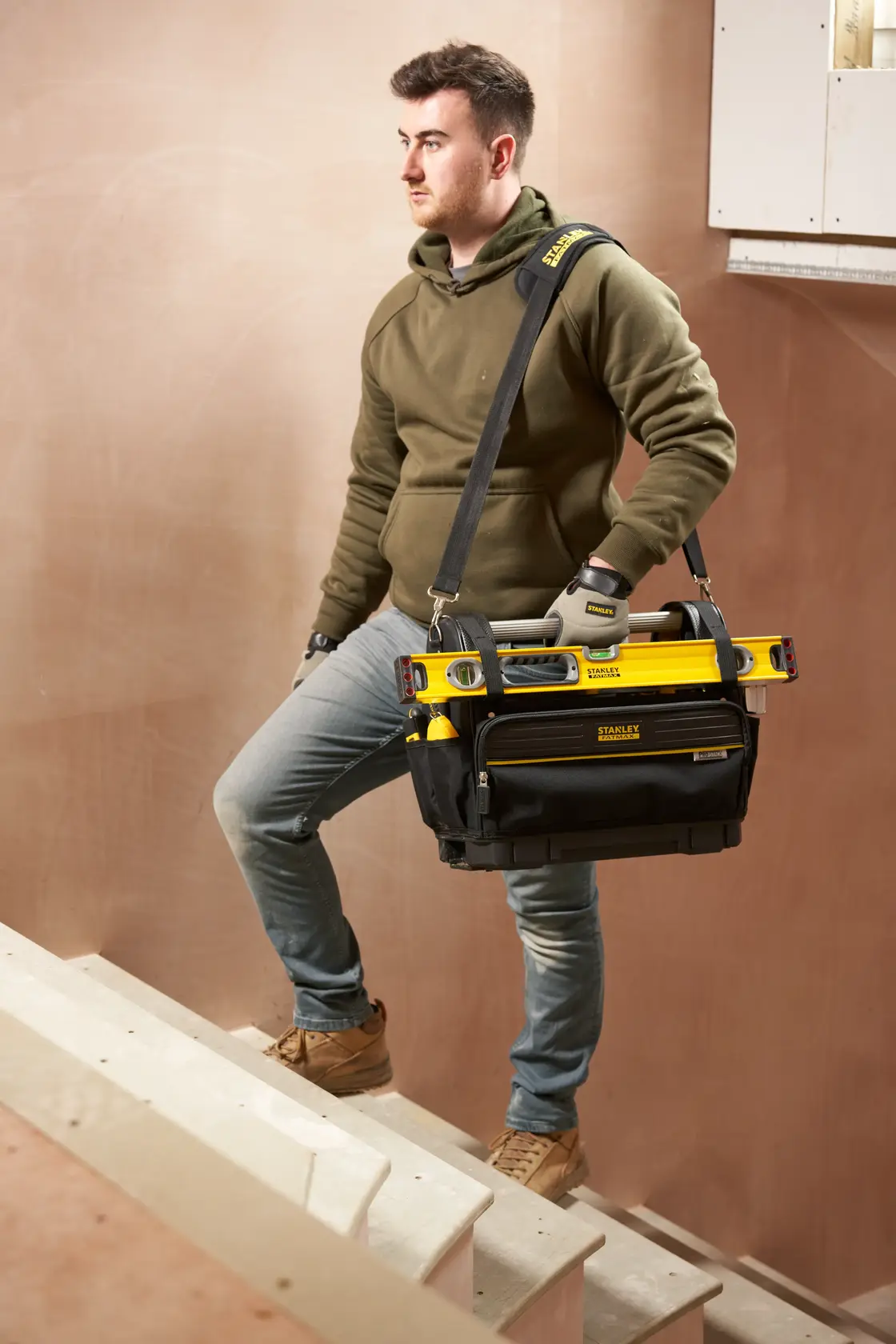 Stanley FMST17626-1 Werkzeugtasche mit gelber Wasserwaage, getragen von Arbeiter auf Treppe, Seitenansicht.