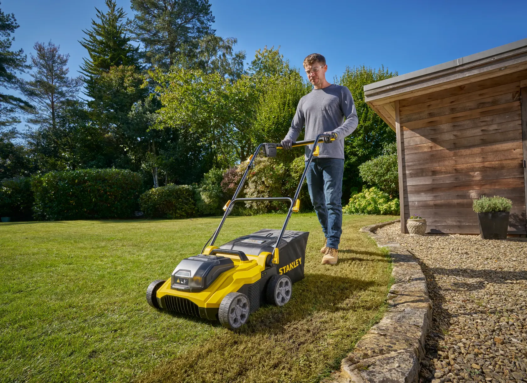Foto des STANLEY SCOEP3801M2 Rasenlüfters auf Gras bei einem Holzschuppen, Person schiebt das Gerät im Freien.