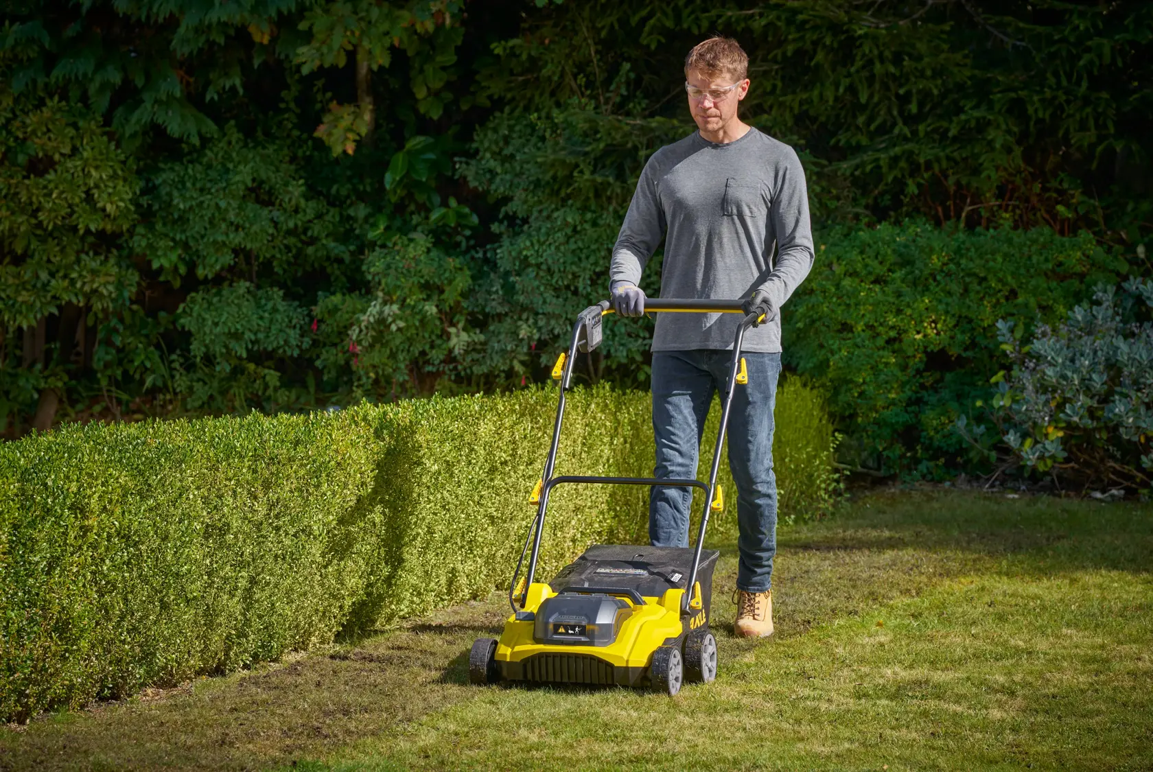 STANLEY SCOEP3801N gelber Vertikutierer im Garten im Einsatz, mit grüner Umgebung im Hintergrund.