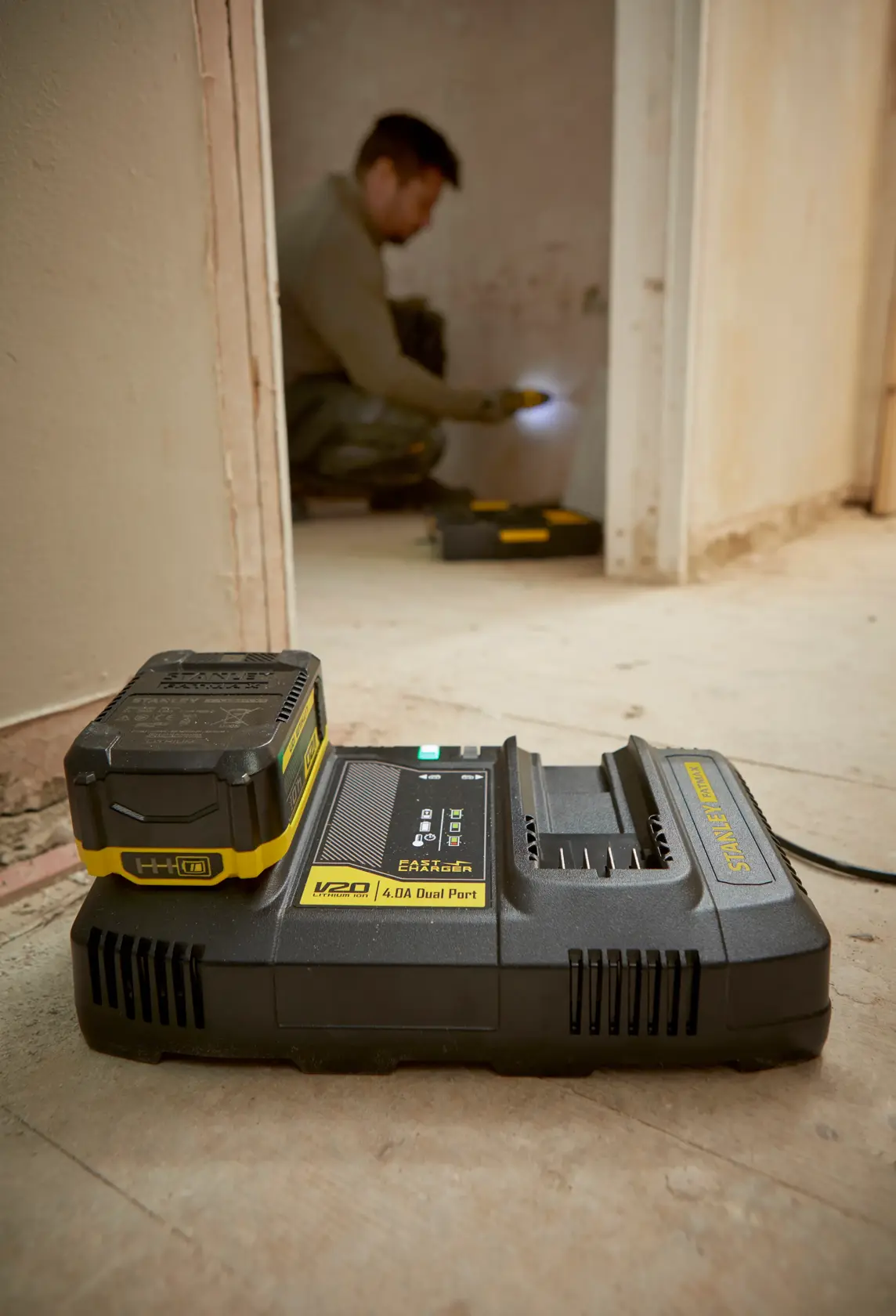 STANLEY SFMCB24 V20 Doppelladegerät mit eingesetztem Akku auf Werkstattboden, Arbeiter im Hintergrund.