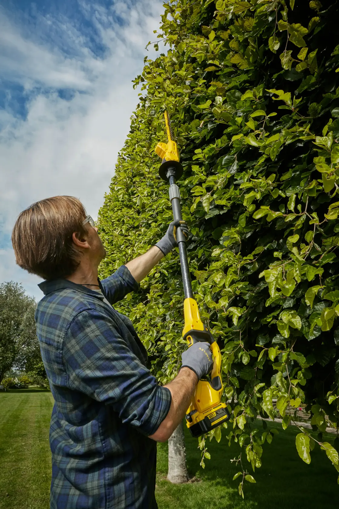 Foto des STANLEY SFMCPH845B Teleskop-Heckenschneiders beim Schneiden hoher grüner Hecken im Freien.