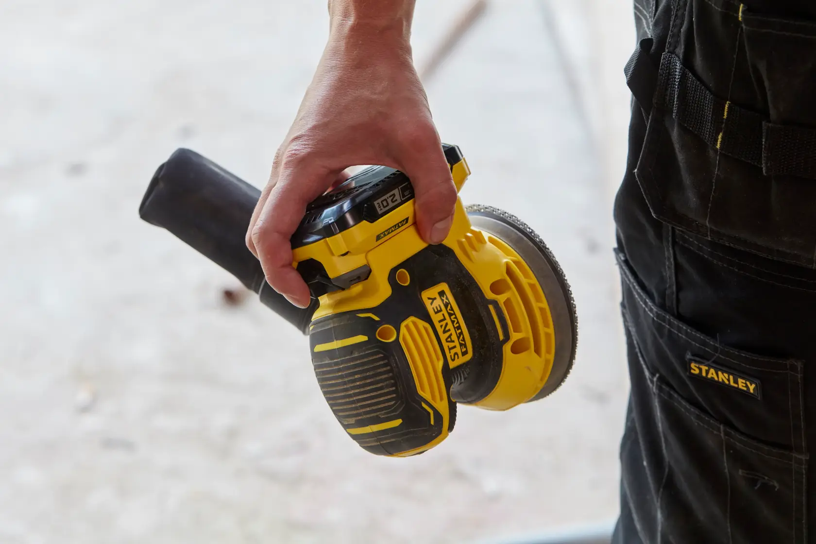 Foto der kabellosen STANLEY SFMCW220B Exzenterschleifer in der Hand, gelb-schwarzes Gehäuse, Arbeitshose sichtbar.