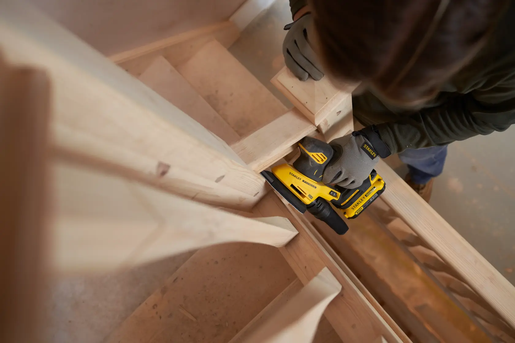 Foto eines Arbeiters mit STANLEY SFMCW221B Schleifer an einer Holztreppe, mit Handschuhen, Innenbereich.