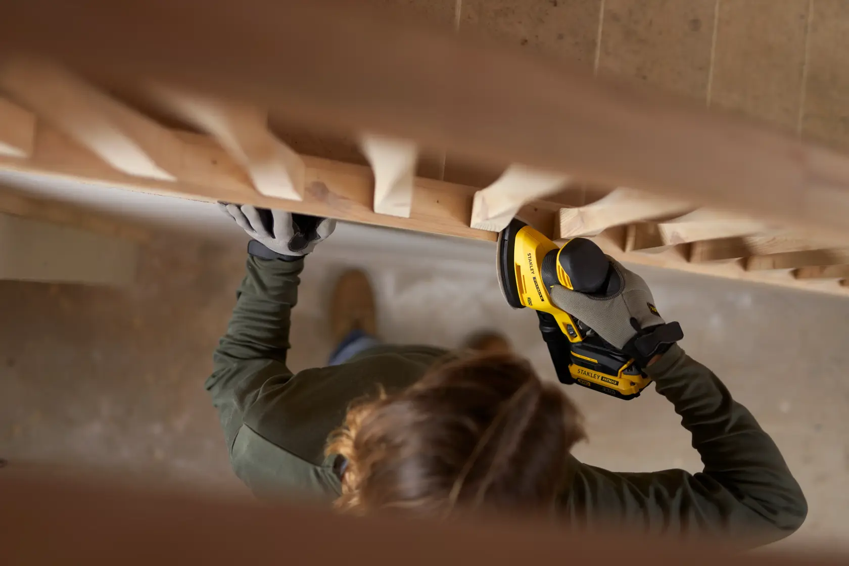 Foto einer Person, die den STANLEY SFMCW221D1S Akku-Schleifer an einem Holzgeländer mit Handschuhen benutzt.