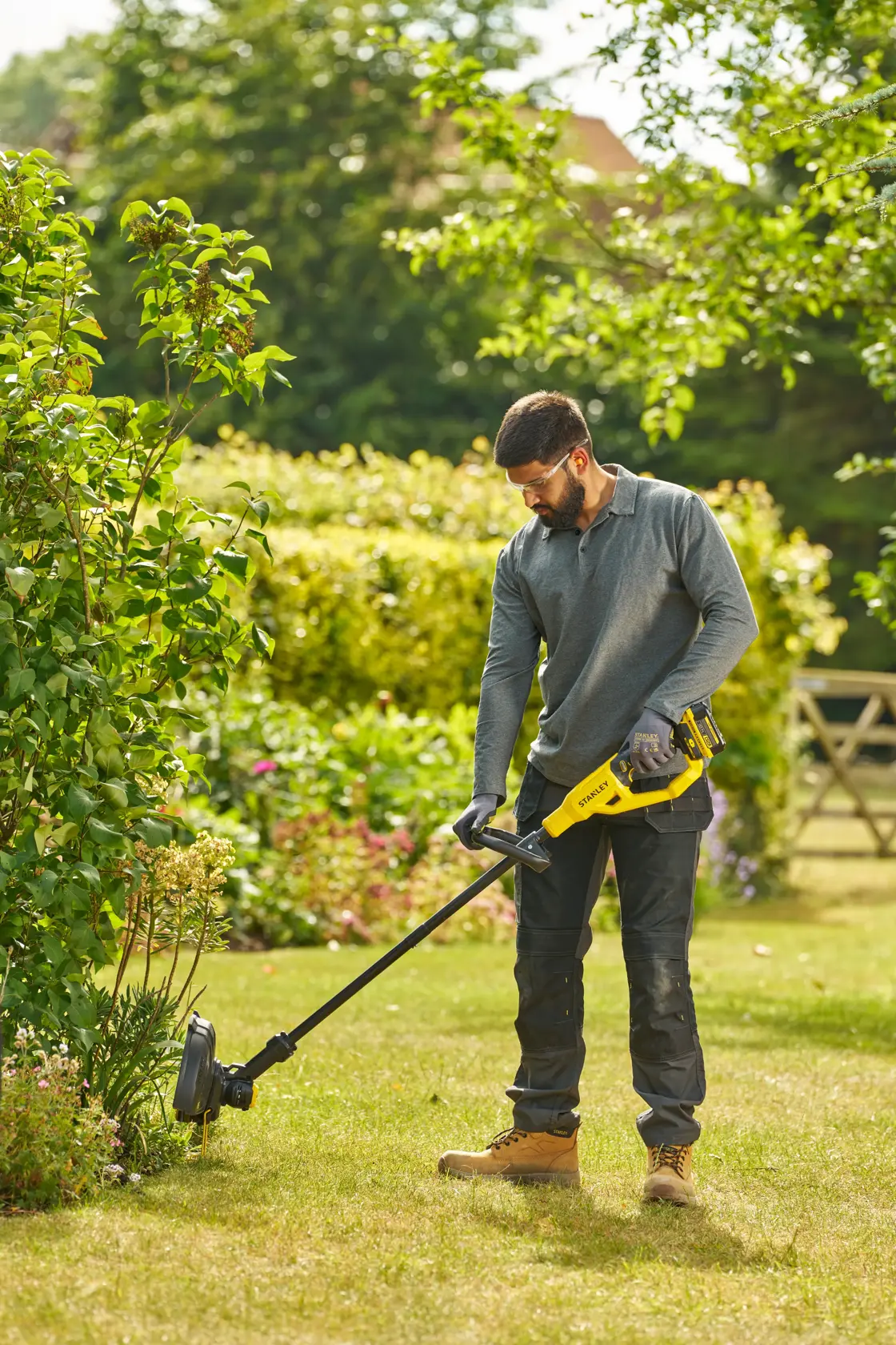 Foto einer Person mit einem STANLEY STCST933B Rasentrimmer im Garten, hält das gelb-schwarze Gerät auf grünem Rasen.