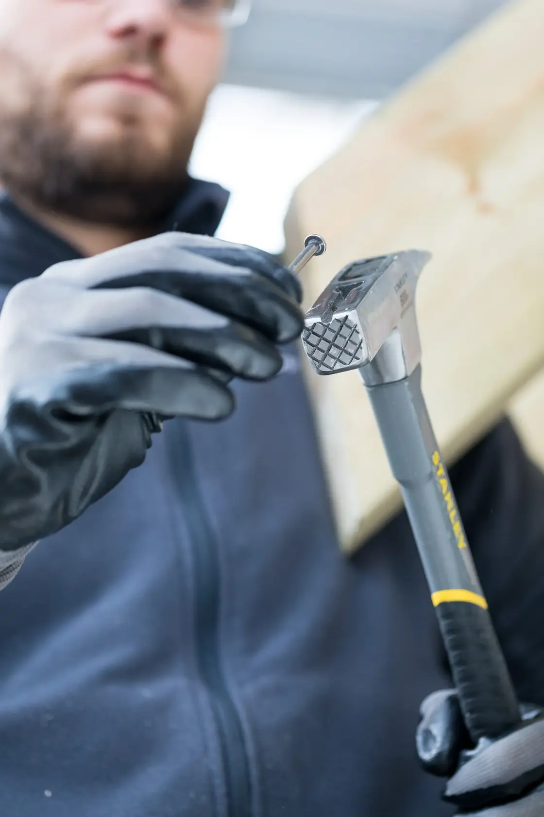 STANLEY STHT0-51312 Hammer Nahaufnahme, behandschuhte Hände halten Nagel und Holz im Hintergrund.