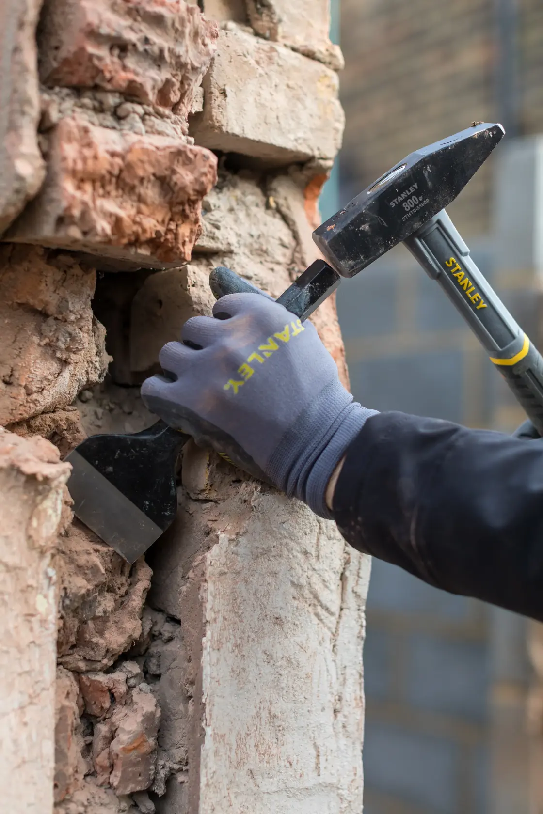 STANLEY STHT0-51909 Fotografie einer behandschuhten Hand, die mit Hammer und Meißel alten Mörtel aus einer Ziegelwand entfernt.