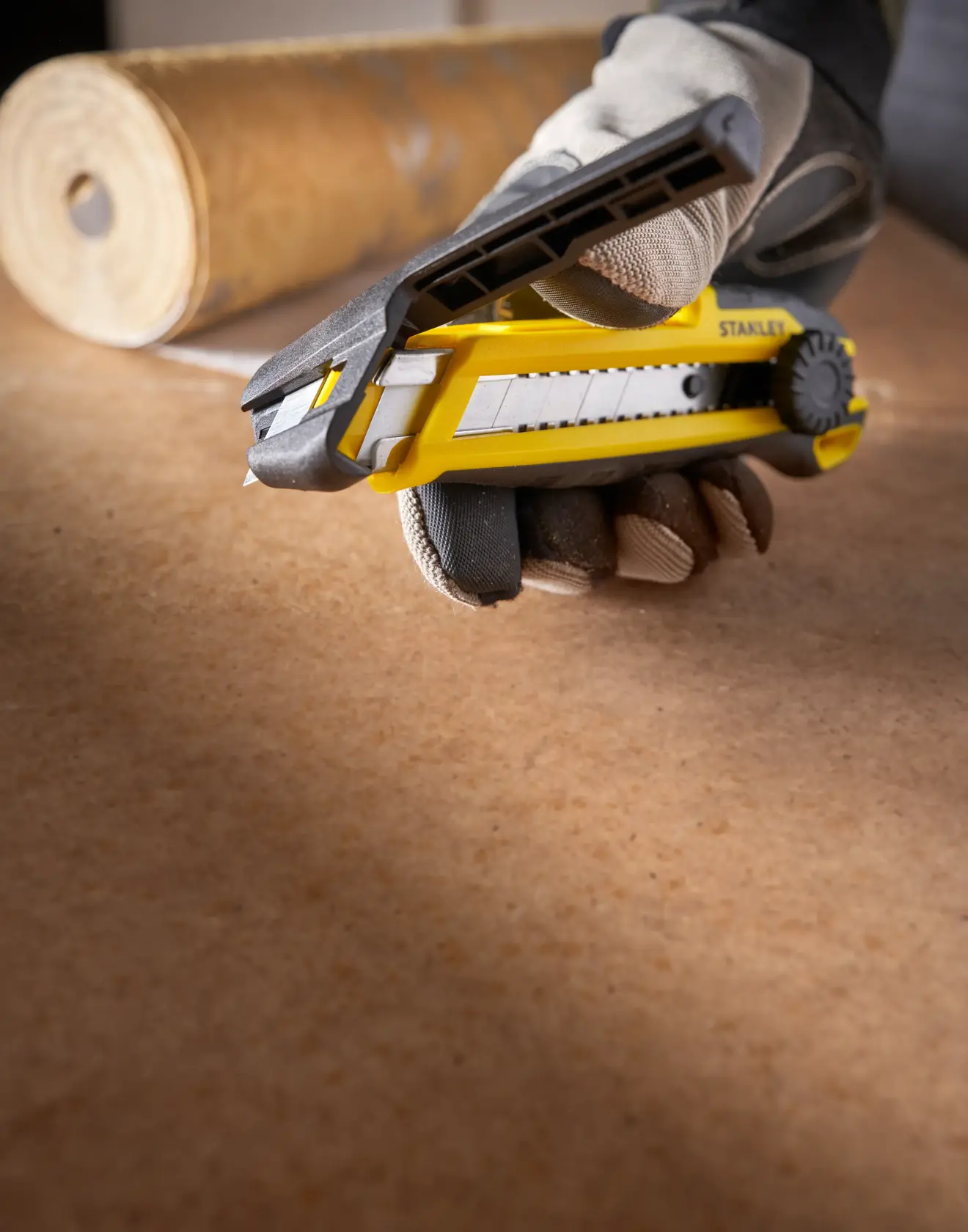 STANLEY STHT10500-0 Cuttermesser in behandschuhter Hand auf Arbeitsfläche, Rolle im Hintergrund, Nahaufnahme.