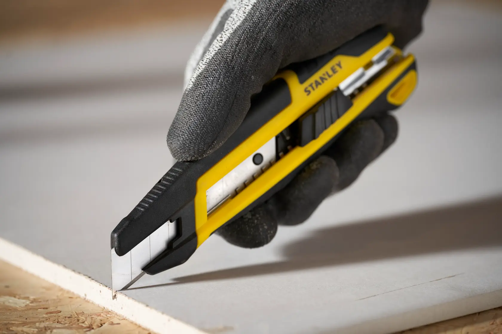 Foto vom STANLEY STHT10500-0 Universalmesser mit schwarz-gelbem Griff, Handschuhhand schneidet Gipskarton.
