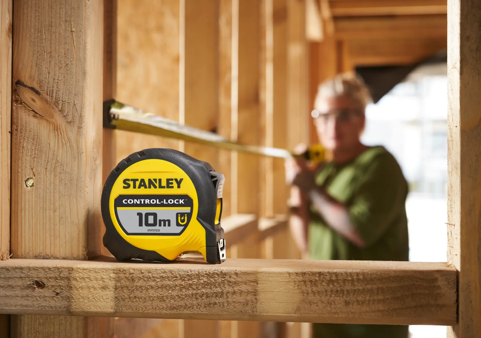 STANLEY STHT37233-0 Bandmaß 10m auf Holzbalken, unscharfe Person misst im Hintergrund.