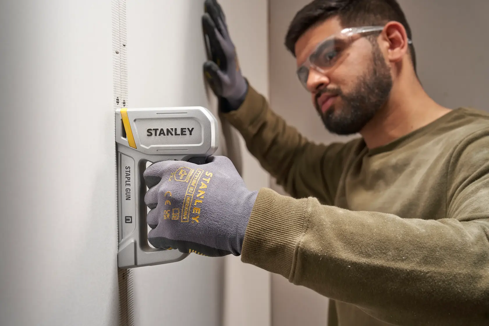 Stanley STHT70150-0 Tacker im Einsatz an Trockenbauwand mit Handschuh, Seitenansicht, Fotoprodukt.