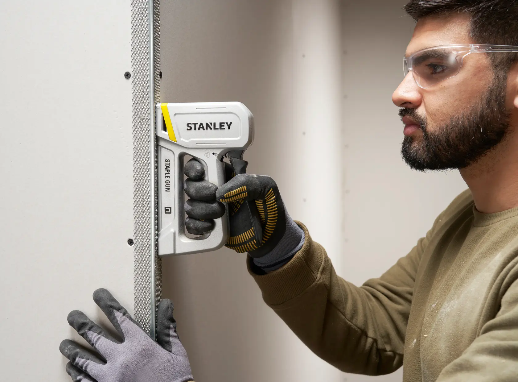 Stanley STHT70150-0 Tacker von Person mit Handschuhen gehalten, befestigt Metalleckprofil an Trockenbauwand.