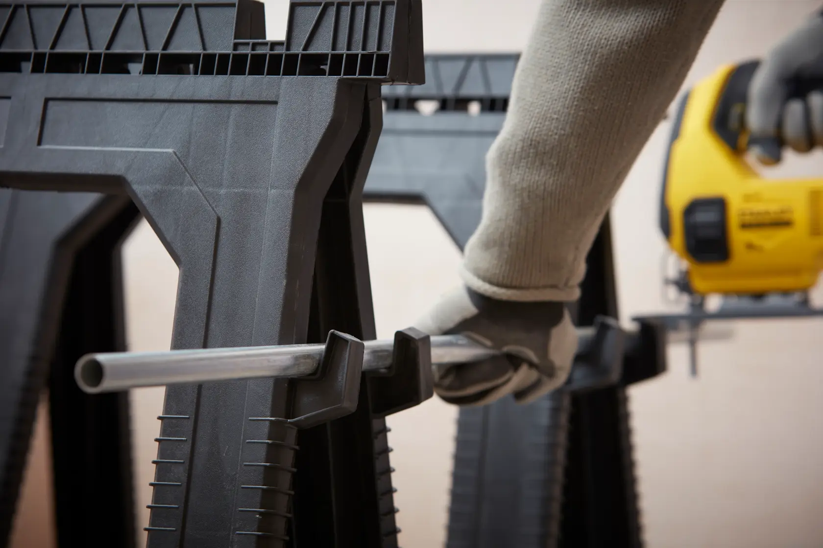 STANLEY STST1-70713 Sägebock Nahaufnahme mit behandschuhter Hand, Metallrohr haltend, DEWALT Stichsäge im Hintergrund.